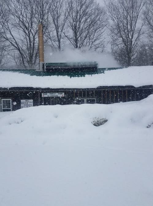 A snowy scene of a rustic building with a chimney, surrounded by deep snow and falling snowflakes.