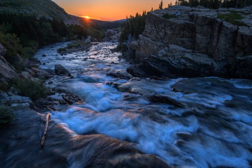 A serene river flows through rocky terrain at sunset, with vibrant colors reflecting off the water and trees in the background.