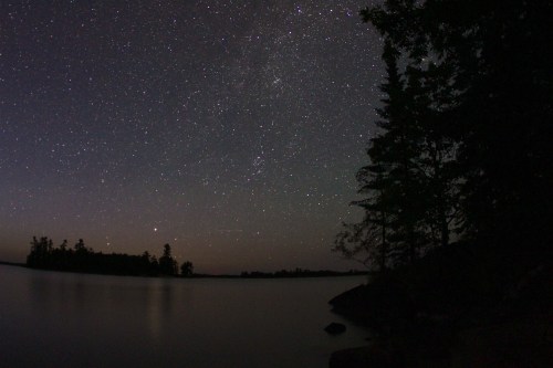 A serene night sky filled with stars over a calm lake, framed by silhouettes of trees on the shore.
