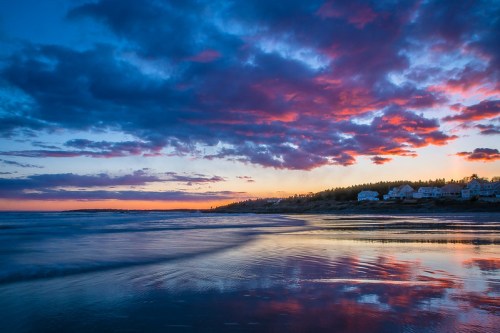 A vibrant sunset over a beach, with colorful clouds reflecting on the water and silhouettes of houses in the background.