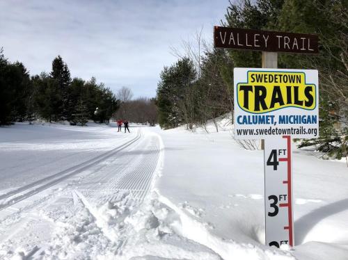 Sign for Valley Trail at Swedetown Trails in Calumet, Michigan, with snow-covered path and trees in the background.