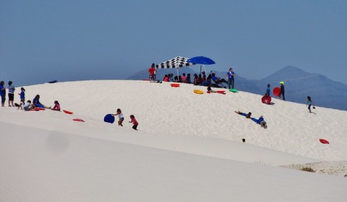 Children playing on a sandy hill, sliding down with colorful sleds, under a checkered umbrella on a sunny day.
