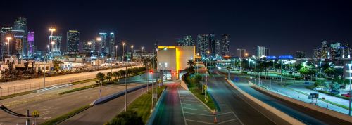 A panoramic view of a city skyline at night, featuring illuminated buildings and empty roads.