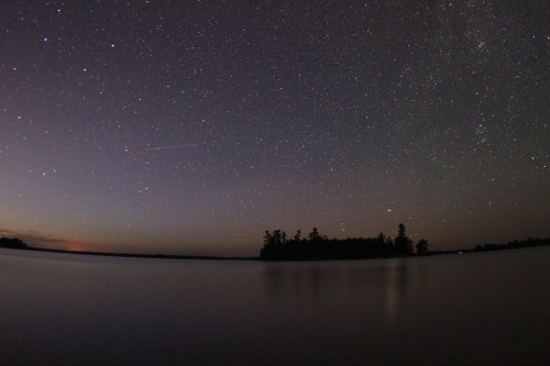A serene night sky filled with stars over a calm lake, with a silhouette of trees on the horizon.