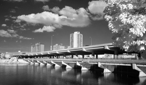 Black and white image of a bridge over a river, with city buildings and clouds in the background.