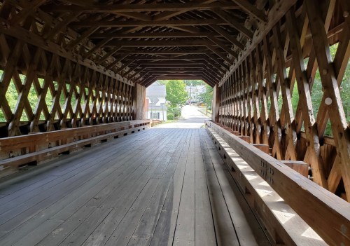 A view inside a wooden covered bridge, showcasing its lattice structure and wooden flooring, leading to a bright exit.