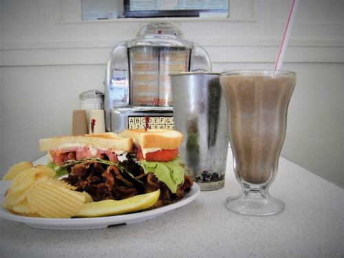 A plate with a sandwich, chips, and a lemon slice next to a milkshake in a retro diner setting.