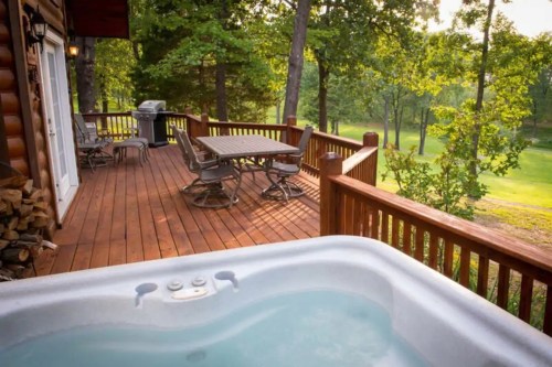 A wooden deck with a hot tub, dining table, and grill, surrounded by trees and a green landscape.