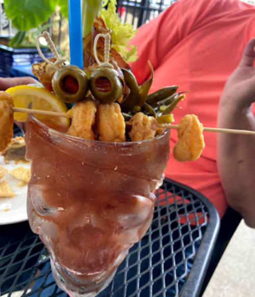 A skull-shaped glass filled with a cocktail, garnished with shrimp, olives, lemon, and pickled vegetables.