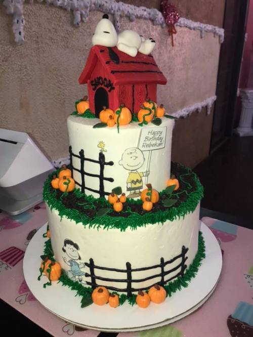 A two-tiered birthday cake decorated with Snoopy on a doghouse, pumpkins, and Peanuts characters.