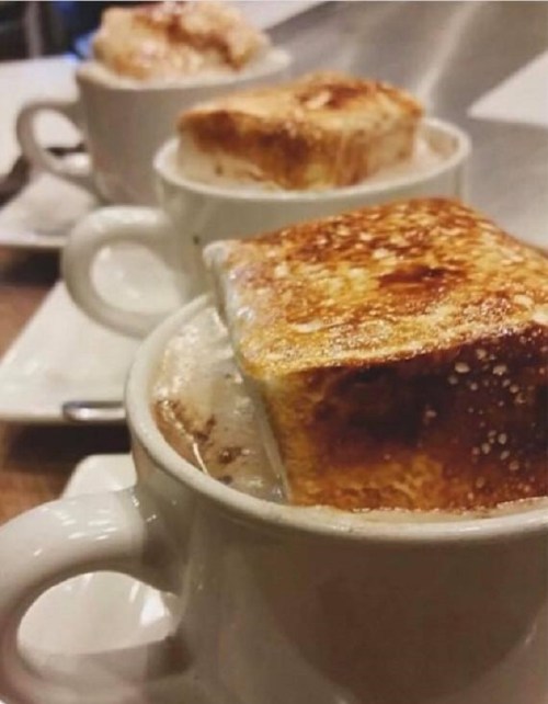 Three cups of hot chocolate topped with toasted marshmallow squares, served on white plates.