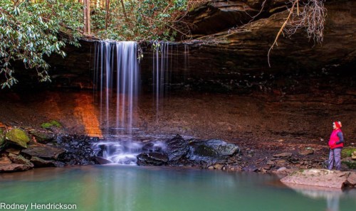 A serene waterfall cascades into a tranquil pool, with a person standing nearby, surrounded by lush greenery.