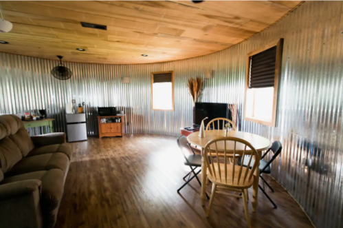 Cozy interior of a round room with corrugated metal walls, featuring a sofa, dining table, and kitchenette.