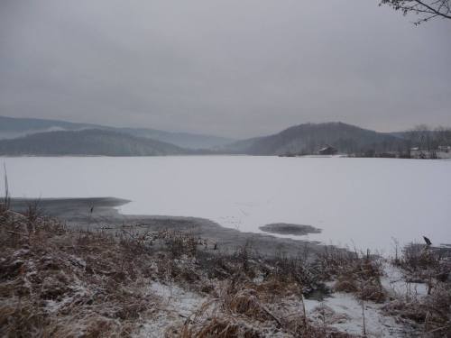 A snowy landscape with a frozen lake, surrounded by hills under a cloudy sky.