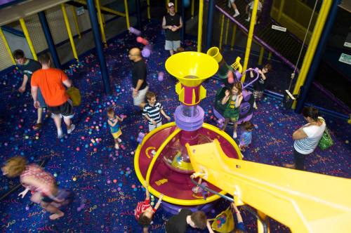 A colorful indoor play area with a large water slide, children playing, and adults supervising in a vibrant, ball-filled space.