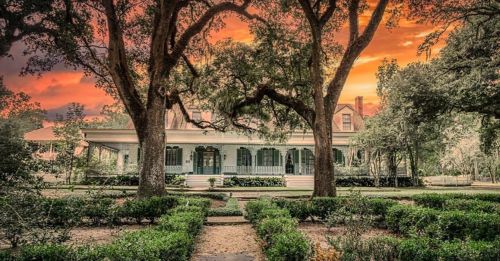 A historic house surrounded by lush greenery and trees, set against a vibrant sunset sky.