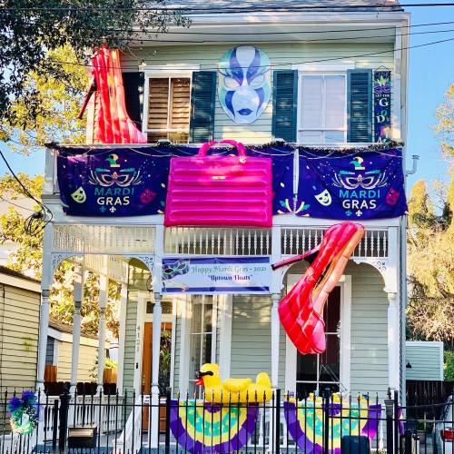 A festive house decorated for Mardi Gras with colorful banners, inflatable shoes, and a large pink suitcase.
