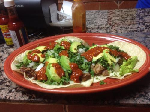 A plate of tacos topped with avocado, cilantro, onions, and red sauce, served on a red dish with condiments in the background.
