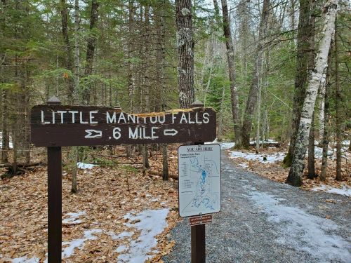 A wooden sign points to Little Manitou Falls, indicating a 0.6-mile trail, surrounded by trees and fallen leaves.