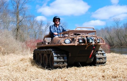 A person driving an all-terrain vehicle on a grassy area near a river, with trees and blue sky in the background.