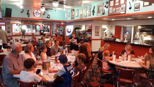A lively diner scene with families and friends gathered around tables, enjoying food and conversation in a retro setting.