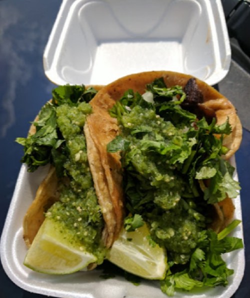 Two tacos filled with green salsa, fresh cilantro, and lime wedges in a takeout container.