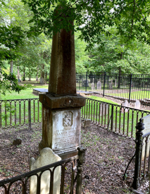 A weathered gravestone with an obelisk shape, surrounded by a wrought iron fence in a lush, green cemetery.