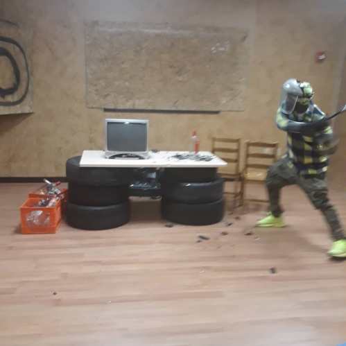 A person in protective gear swings a bat near a makeshift table made of tires and a monitor in a chaotic room.