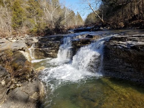 A serene waterfall cascades over rocky ledges into a clear pool, surrounded by trees and a blue sky.