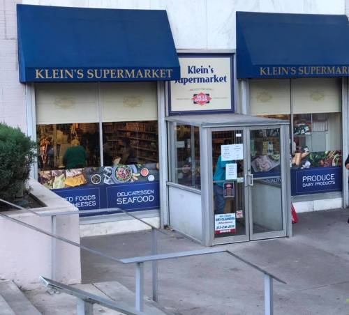 Exterior view of Klein's Supermarket, featuring signs for deli, seafood, and produce, with shoppers visible inside.