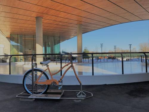 An orange bicycle is parked on a rack near an empty ice skating rink under a modern architectural structure.
