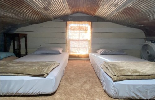 Two unmade beds with blankets in a cozy attic room, featuring a window with natural light and a fan in the corner.