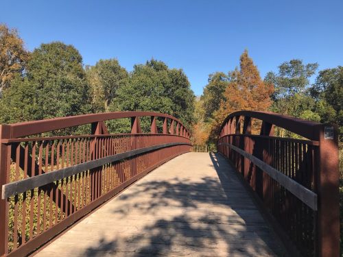 A metal bridge arches over a path, surrounded by trees and a clear blue sky. Autumn colors are visible in the foliage.