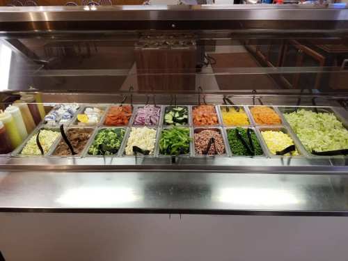 A salad bar featuring various fresh vegetables, toppings, and dressings in clear containers on a stainless steel counter.
