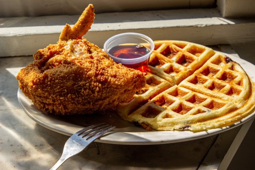 A plate with crispy fried chicken, two waffles, and a small cup of syrup, set near a window.