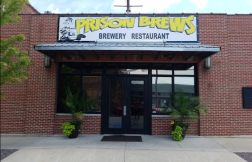 Exterior of Prison Brews brewery and restaurant, featuring a sign and large windows, with potted plants at the entrance.