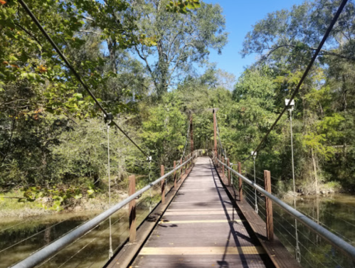 A wooden suspension bridge spans a calm river, surrounded by lush green trees under a clear blue sky.