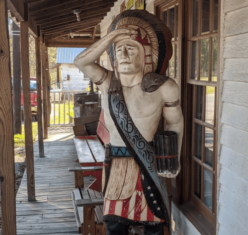 A wooden statue of a Native American figure, wearing a headdress and holding a cup, stands outside a rustic building.