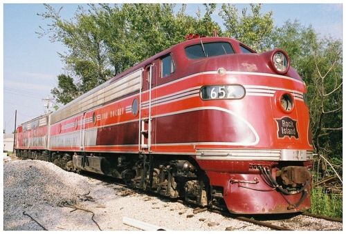 A vintage red and silver train engine parked on a gravel track, surrounded by greenery.