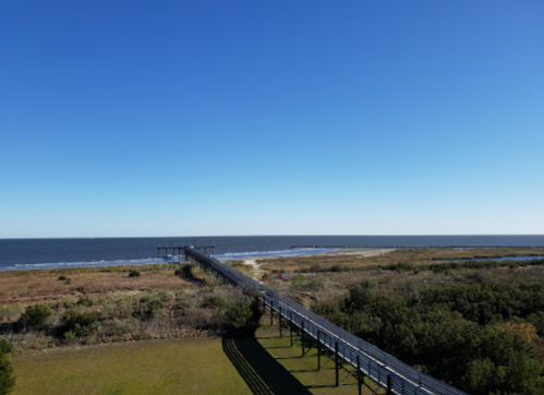 A wooden pier extends into a calm sea under a clear blue sky, surrounded by grassy land and sparse vegetation.
