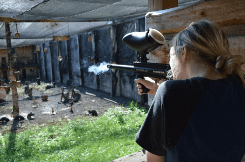 A person aims a paintball gun at a target range, smoke rising from the barrel, surrounded by outdoor scenery.