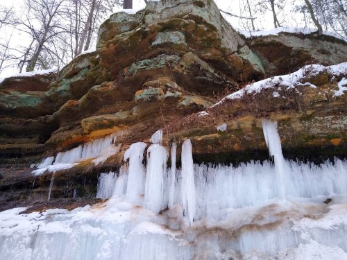 Icicles hang from a rocky cliff, surrounded by snow and trees, showcasing a winter landscape.