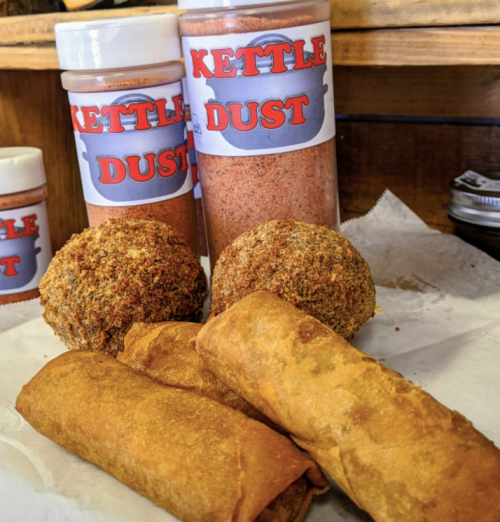 Two crispy spring rolls and two golden-brown fried balls, with jars of "Kettle Dust" seasoning in the background.