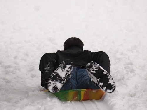 A person sledding down a snowy hill, lying face down on a colorful sled with their feet in the air.