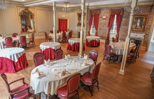 Elegant dining room with round tables, red accents, and vintage decor, featuring chandeliers and brick walls.