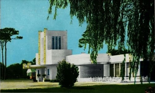 A modernist building with a curved facade, large windows, and a tall tower, surrounded by trees and greenery.