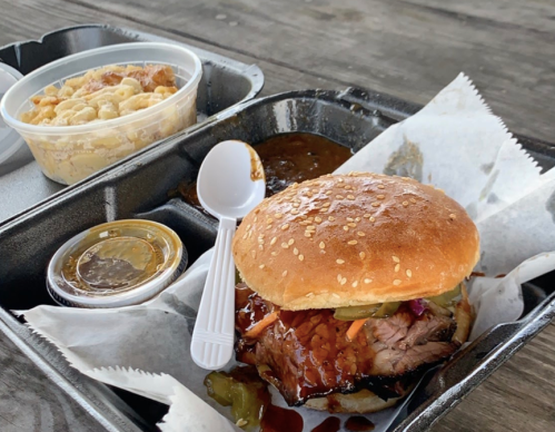 A sesame seed bun sandwich with brisket, pickles, and sauce, served with macaroni and cheese in a takeout container.