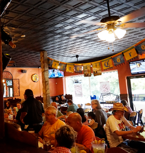 A lively restaurant interior with diners enjoying meals, colorful decorations, and a ceiling fan overhead.