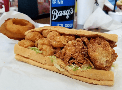 A fried shrimp sandwich with lettuce in a hoagie roll, alongside an onion ring and a Barq's soda.