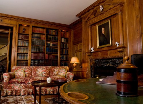 A cozy library with wooden walls, a floral sofa, bookshelves, a fireplace, and a portrait on the wall.